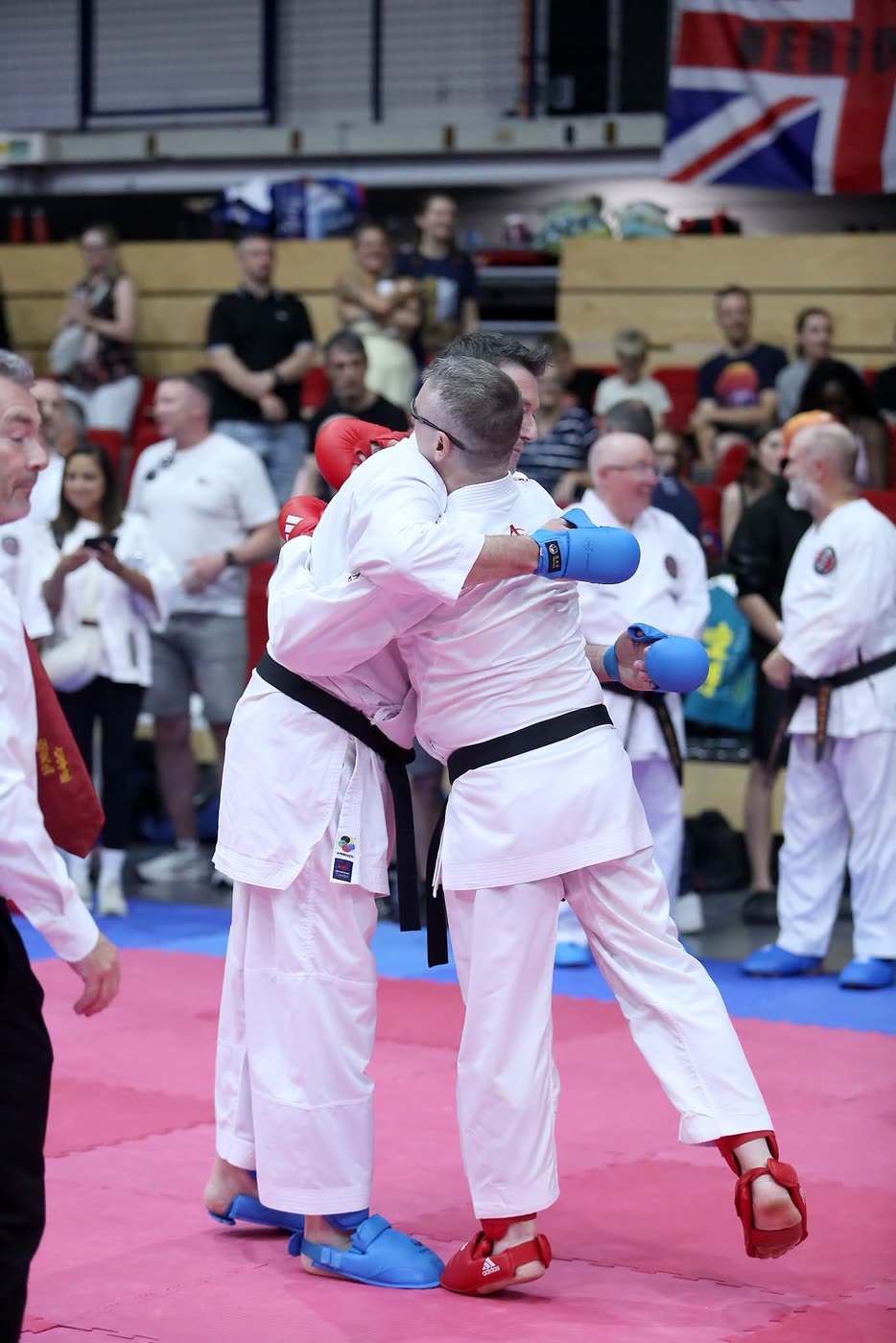 Two senior competitors closing the distance in a kumite bout, the referee watching from the mat edge.