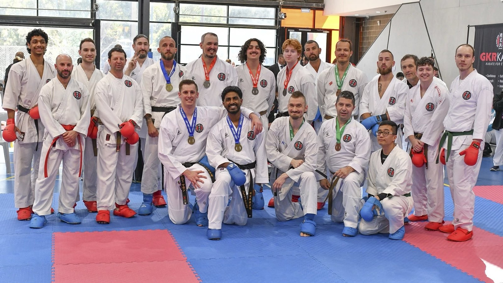 A large group of adult competitors in their gis and sparring gear, many wearing medals, posed together on the mats.