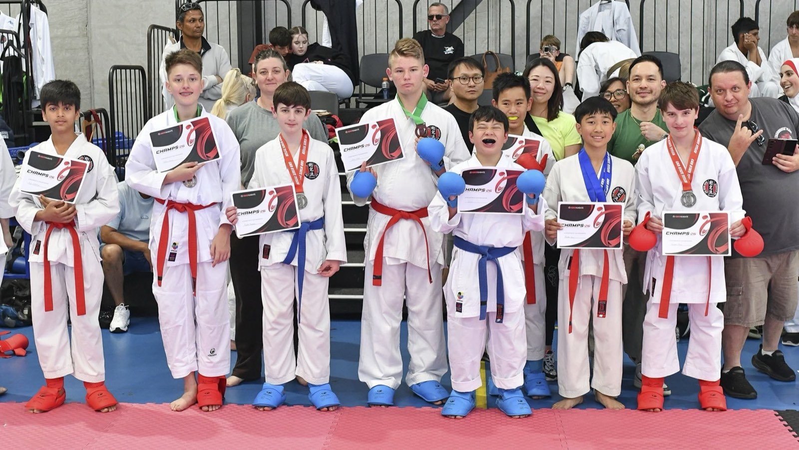 A line of junior competitors holding up their Champs '26 certificates and medals, family members behind them.