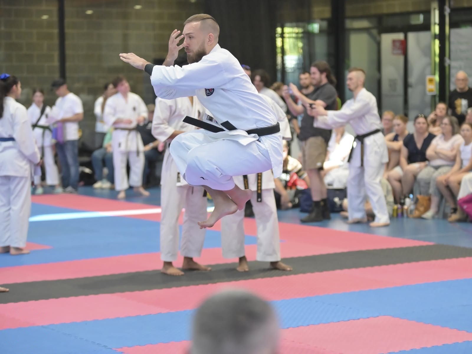 A black belt mid-air during a kata, knees tucked, with competitors and spectators around the mat.