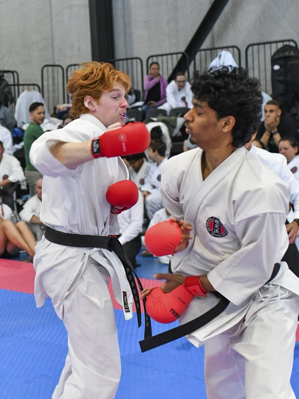 Two senior black belts in red gloves mid-exchange in a kumite bout, spectators watching from the stands.